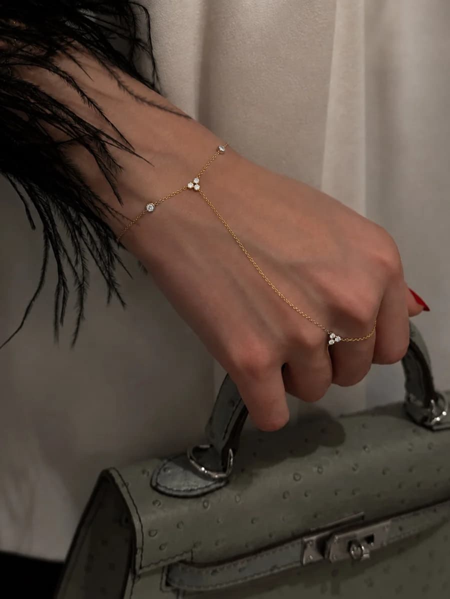 A gold-tone hand chain with tiny crystal-set clusters, worn across the back of a hand holding a sage-green bag.