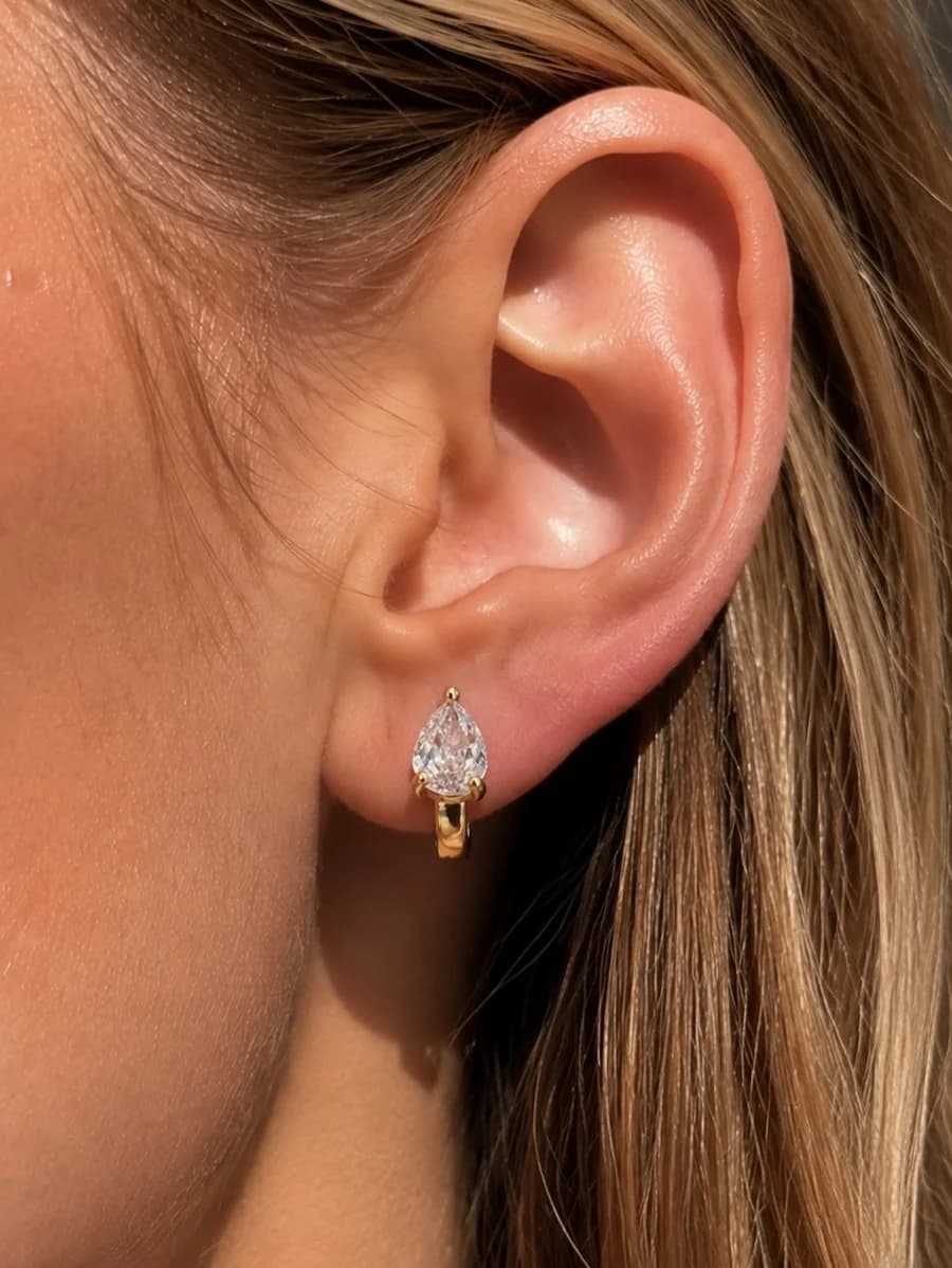 A gold-tone huggie hoop earring with a clear pear-shaped crystal drop, worn on an ear.