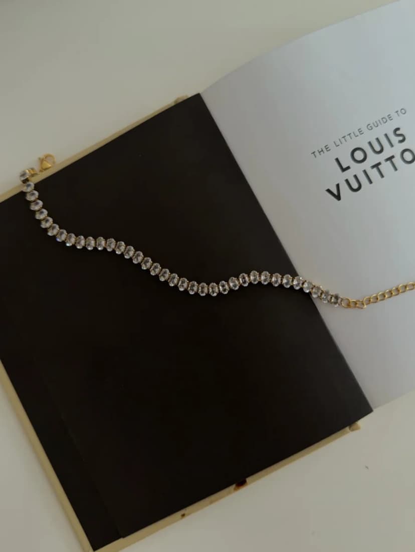 A bracelet with a continuous line of clear oval crystals and a gold-tone chain, laid on an open book.