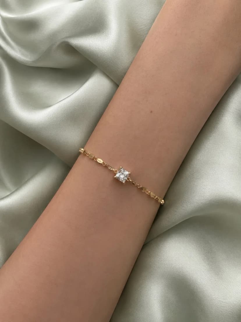 A person's arm wearing a gold-tone bracelet with flat links and a square crystal charm, against pale green silk fabric.