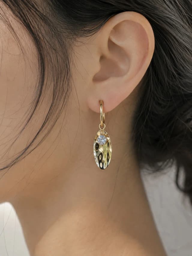 A close-up of a woman's ear wearing a gold-tone hoop earring with an oval, hammered charm and a small blue crystal.