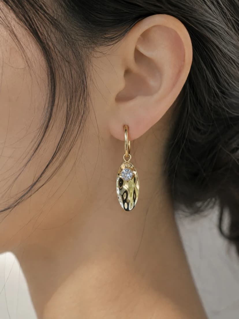 A close-up of a woman's ear wearing a gold-tone hoop earring with an oval, hammered charm and a small blue crystal.