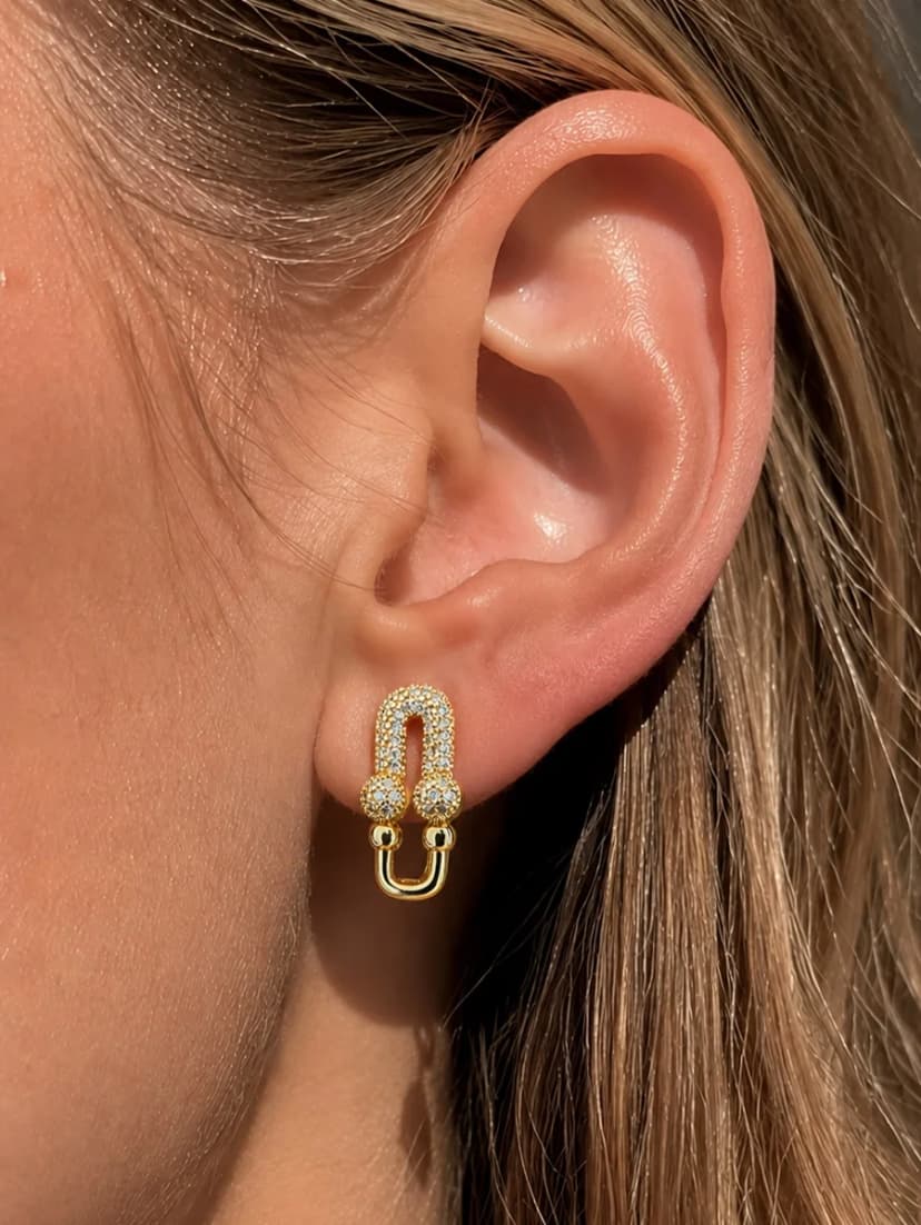 Close-up of a person's ear wearing a gold-tone U-shaped stud earring, adorned with small clear crystals.