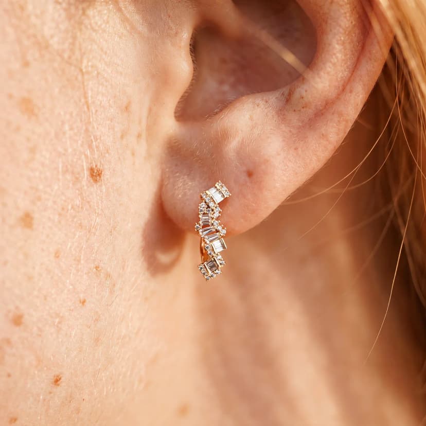 Close-up of a person's ear wearing a small gold-tone stud earring with a curved design, set with various clear crystals.