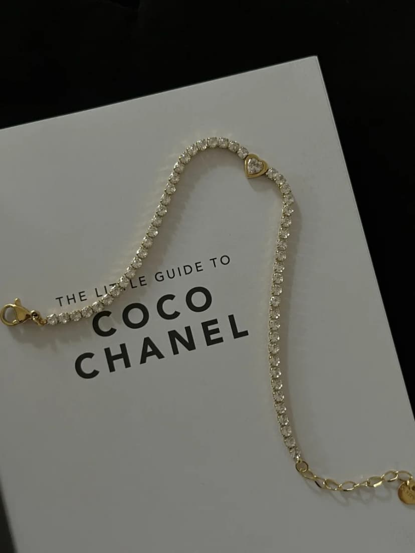 A gold-tone bracelet with a line of clear crystals and a small heart charm, casually draped over a white book.
