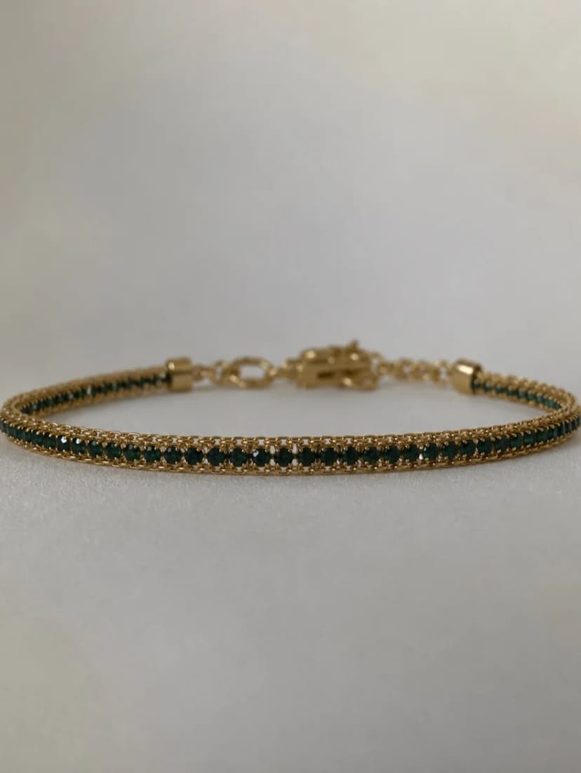 A gold-tone bracelet featuring a delicate chain woven with many tiny, dark green beads, lying on a white surface.