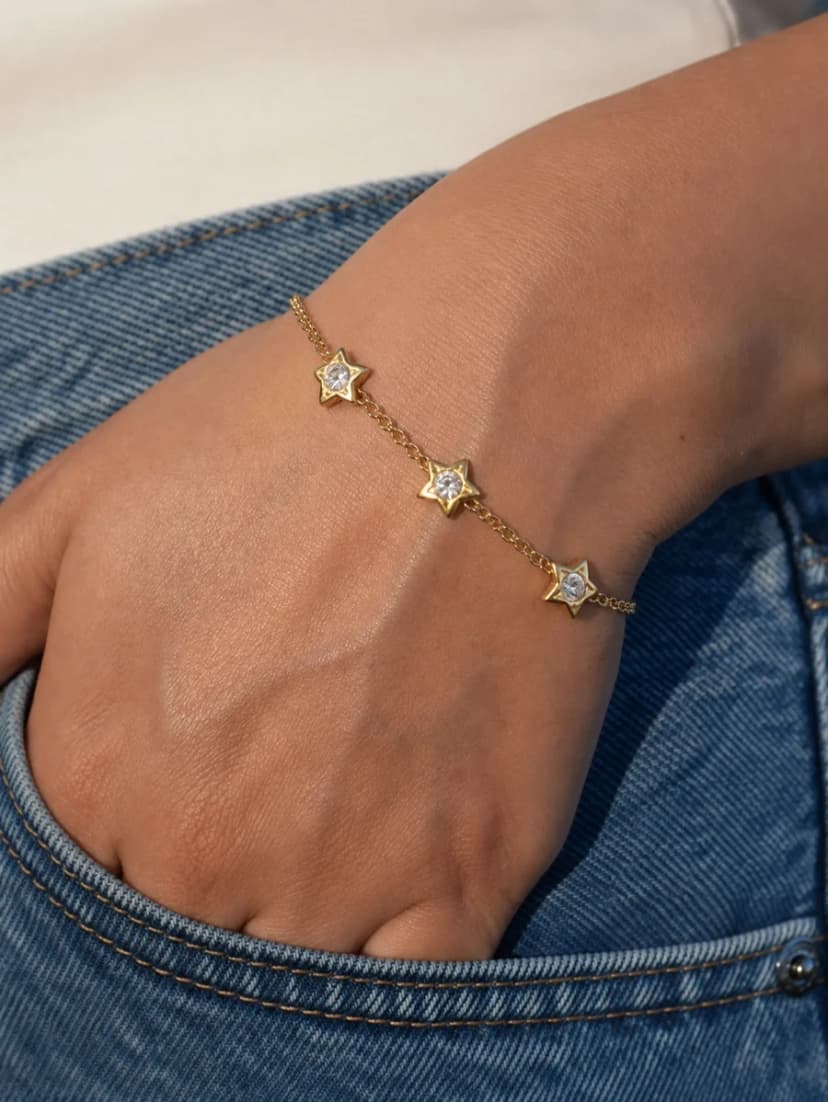 Close-up of a person's wrist wearing a gold-tone bracelet with three crystal-set star charms, resting on blue jeans.