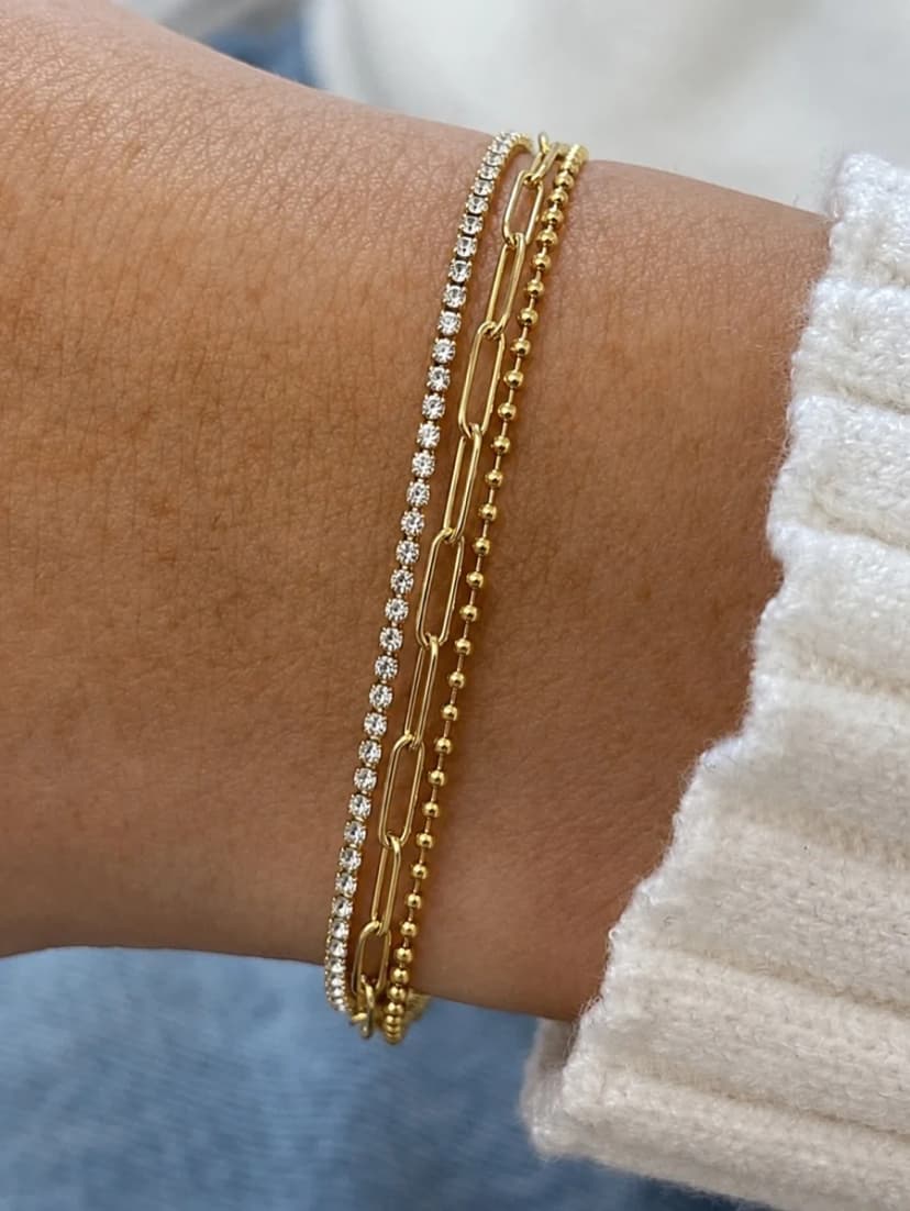 Close-up of a person's wrist wearing three stacked gold-tone bracelets: one crystal-set, one paperclip, one beaded.