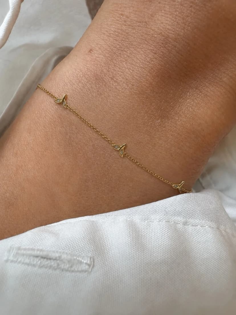 A person's arm wears a delicate gold-tone chain bracelet with small sparkling leaf charms, against white fabric.