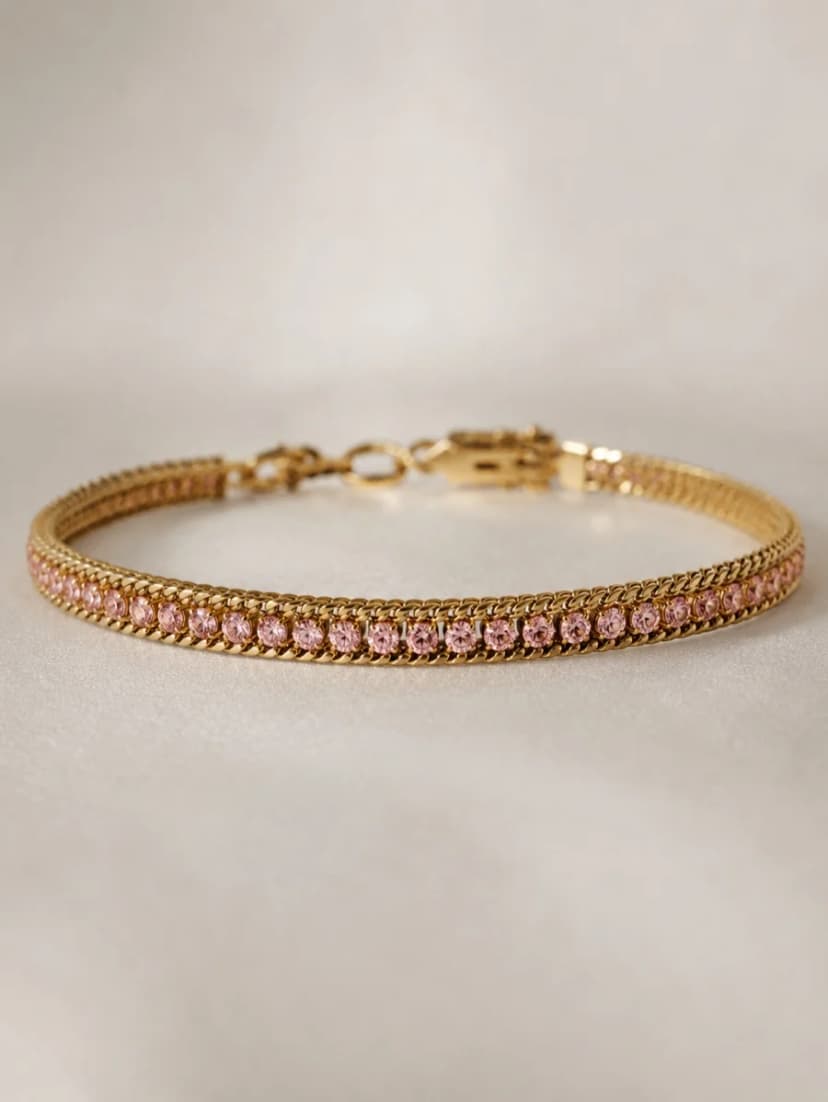 A gold-tone rigid bracelet with a single row of delicate pink crystals set into a textured band, on a light surface.