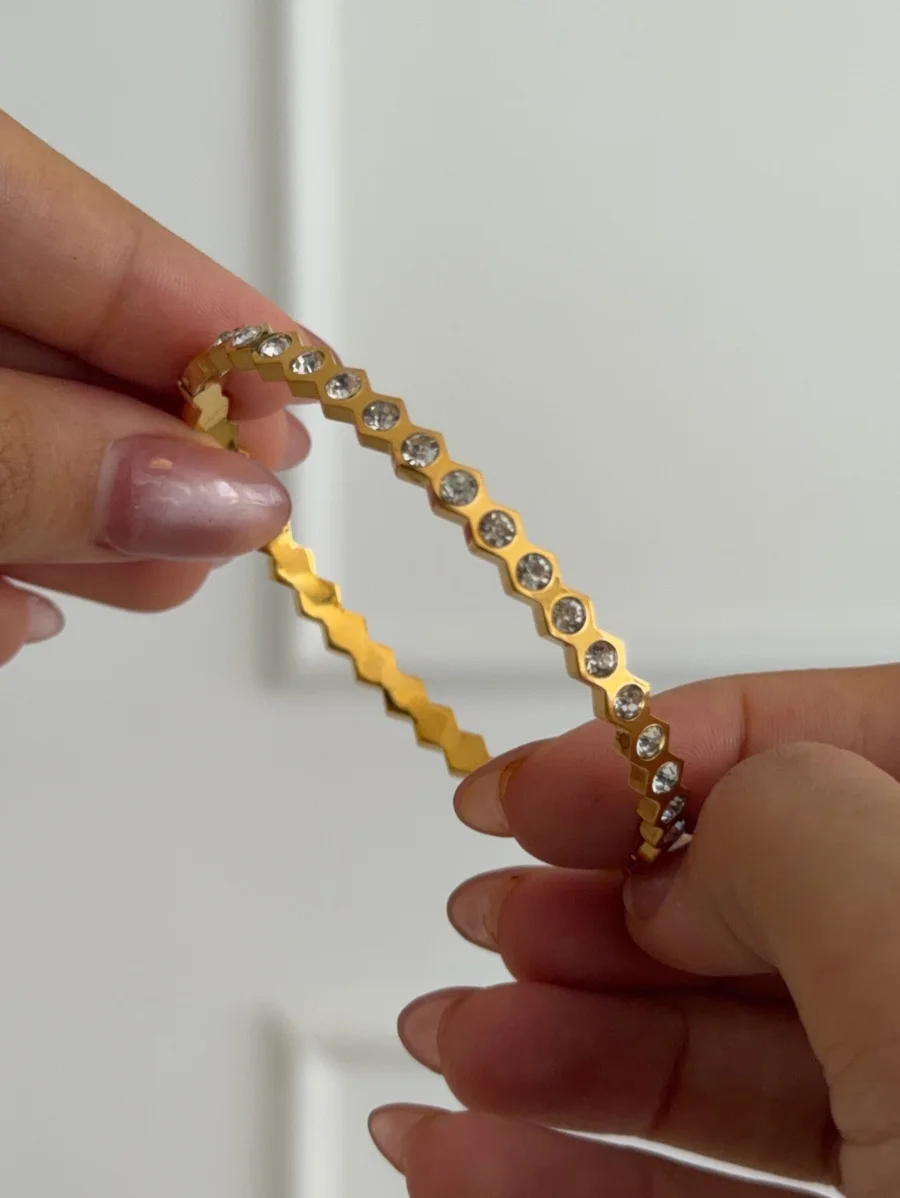Two hands with light pink manicured nails hold a shiny gold-tone bangle with hexagonal crystal-set segments.