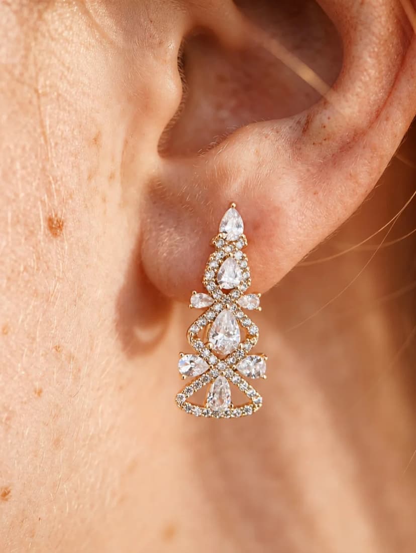 A close-up of an ear wearing a gold-tone drop earring with intricate designs of clear, sparkling pear-shaped and round crystals.