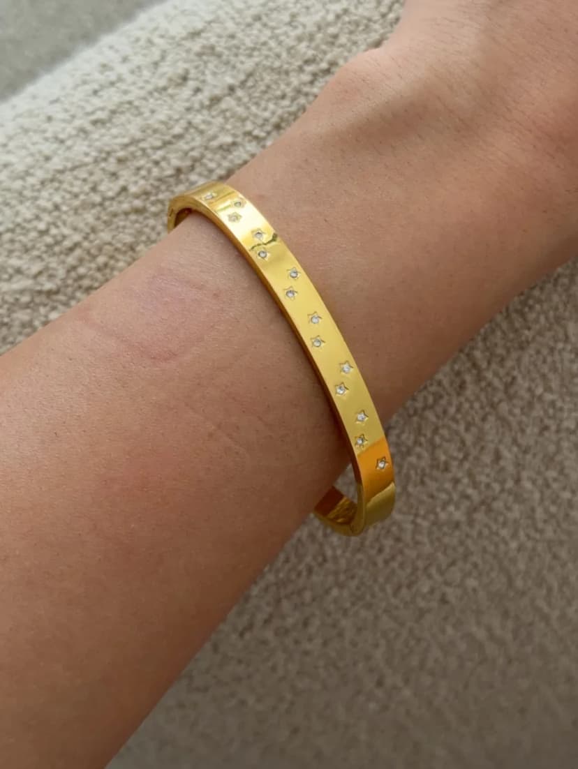 A gold-tone cuff bracelet with scattered clear crystals is worn on a wrist, resting against textured light brown fabric.