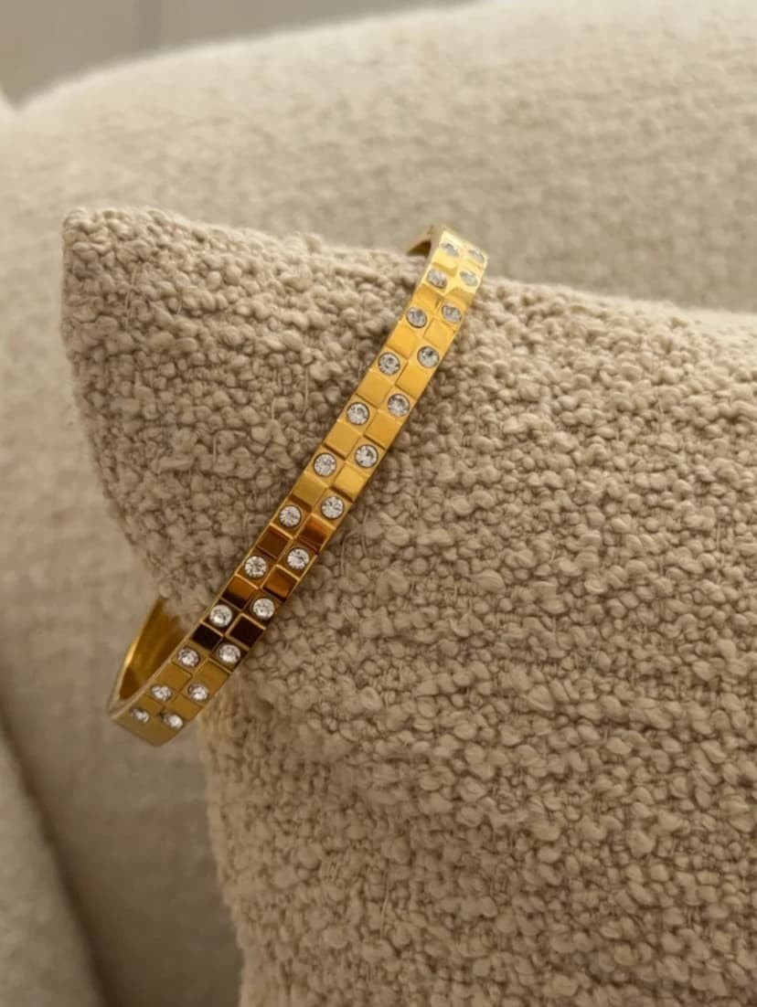 A gold-tone cuff bracelet with two rows of square-set clear crystals rests on a textured light brown cushion.
