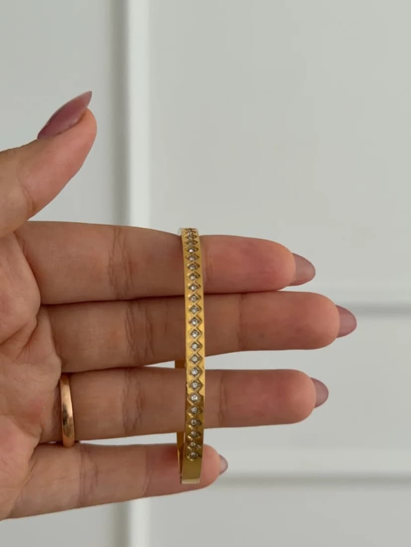 A hand with light pink manicured nails holds a thin gold-tone bangle with a continuous line of tiny clear chevron-set crystals. A simple gold ring is