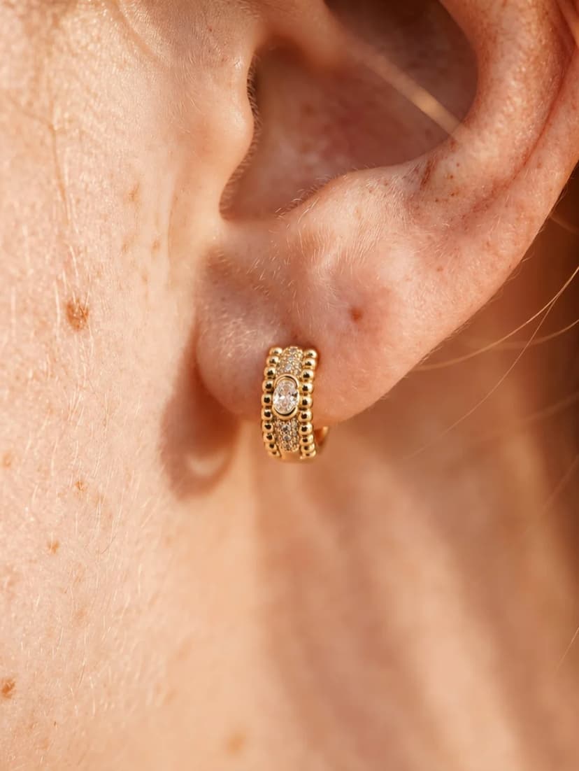 Close-up of an earlobe wearing a small gold-tone hoop earring with beaded edges and a crystal-set oval centre.