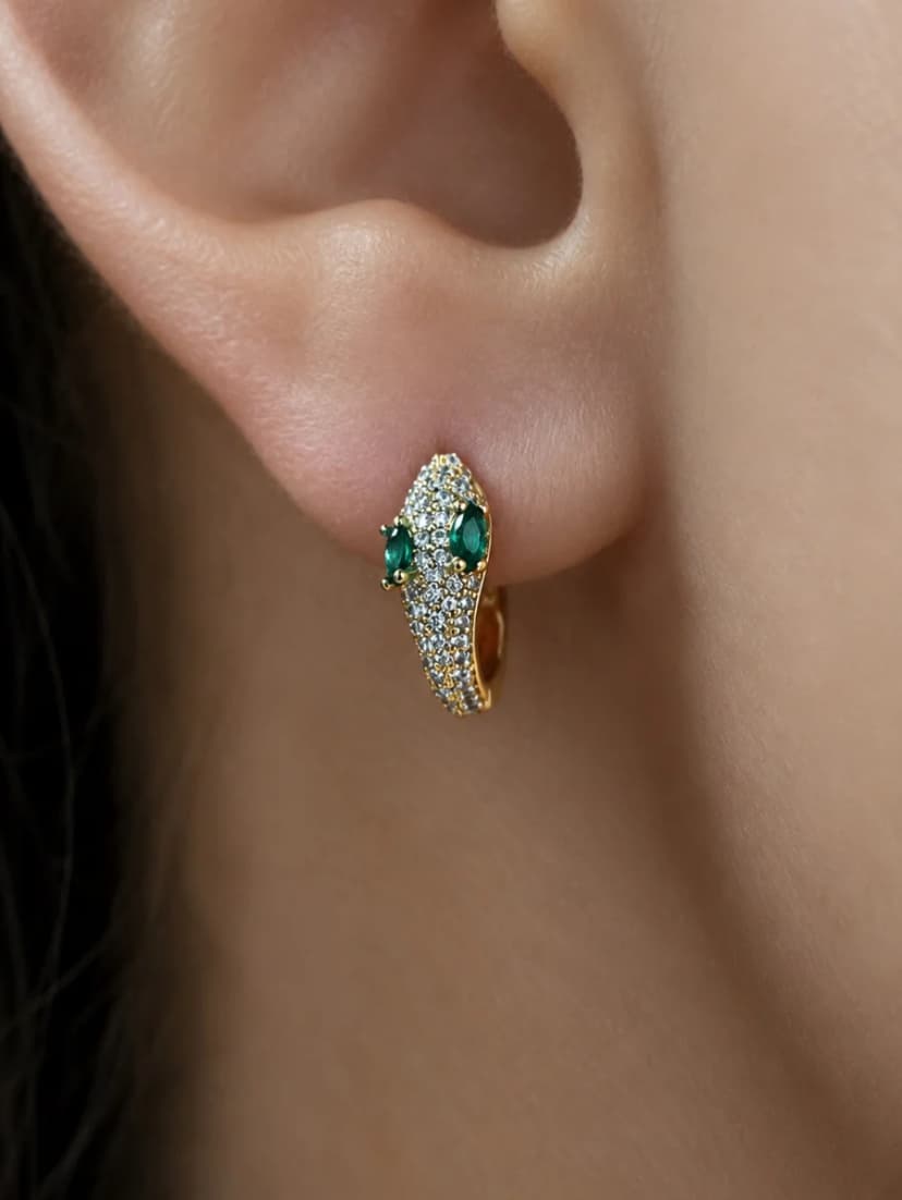 Close-up of a person's earlobe wearing a gold-tone hoop earring shaped like a serpent's head, adorned with clear and green crystals.