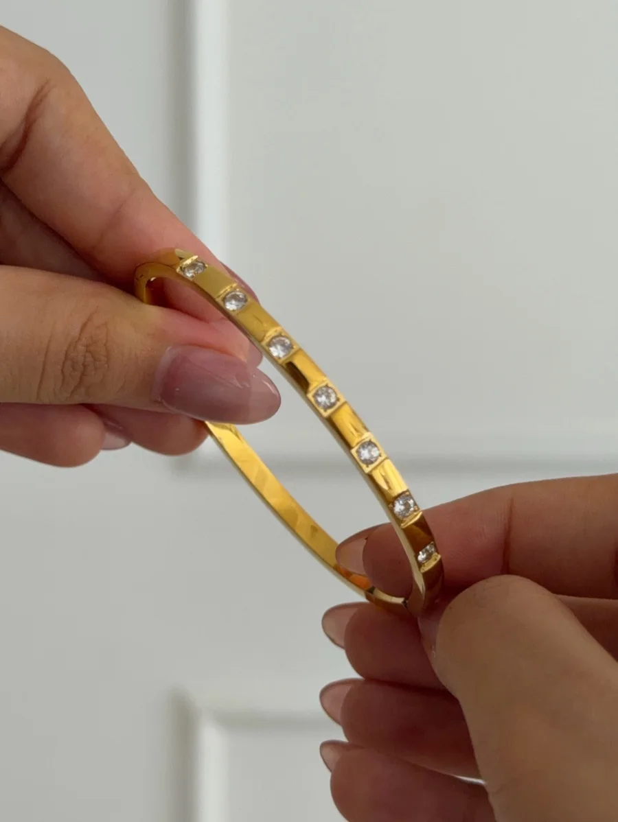 Two hands hold a gold-tone bangle bracelet featuring evenly spaced square-set clear crystals against a white background.