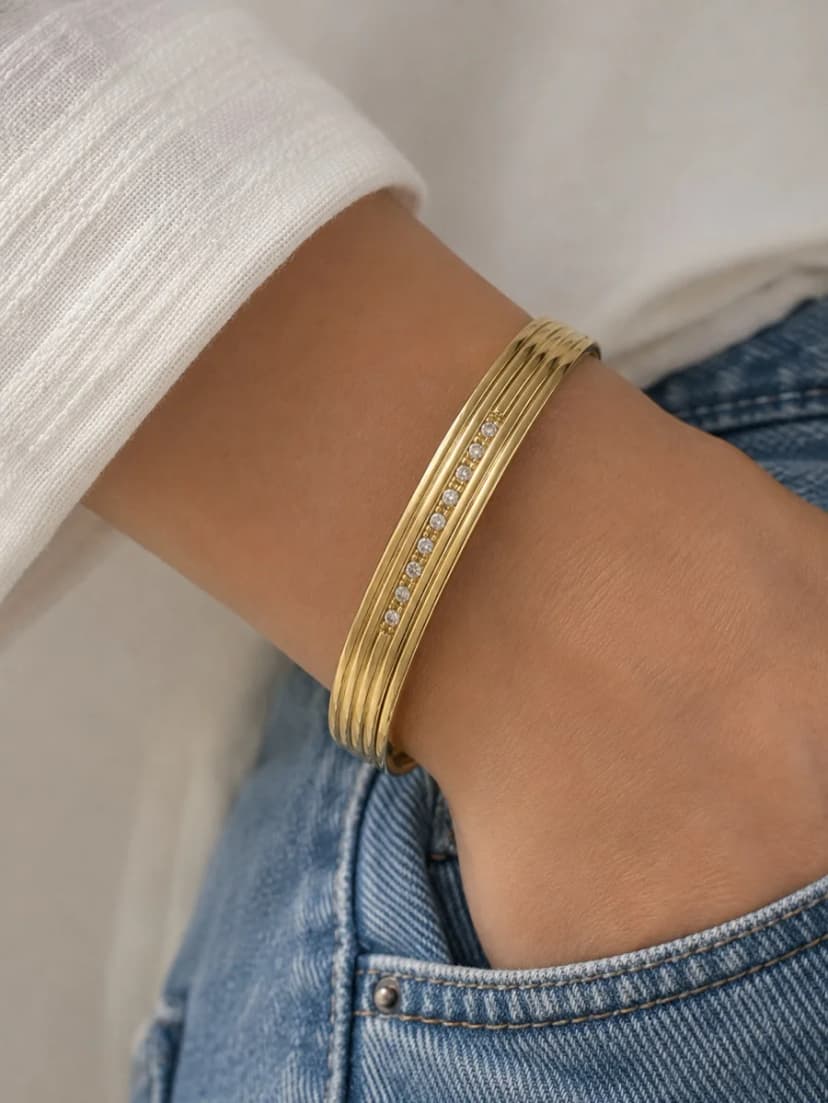 A person's wrist wearing a gold-tone cuff bracelet with parallel ridges and a central line of sparkling crystals.