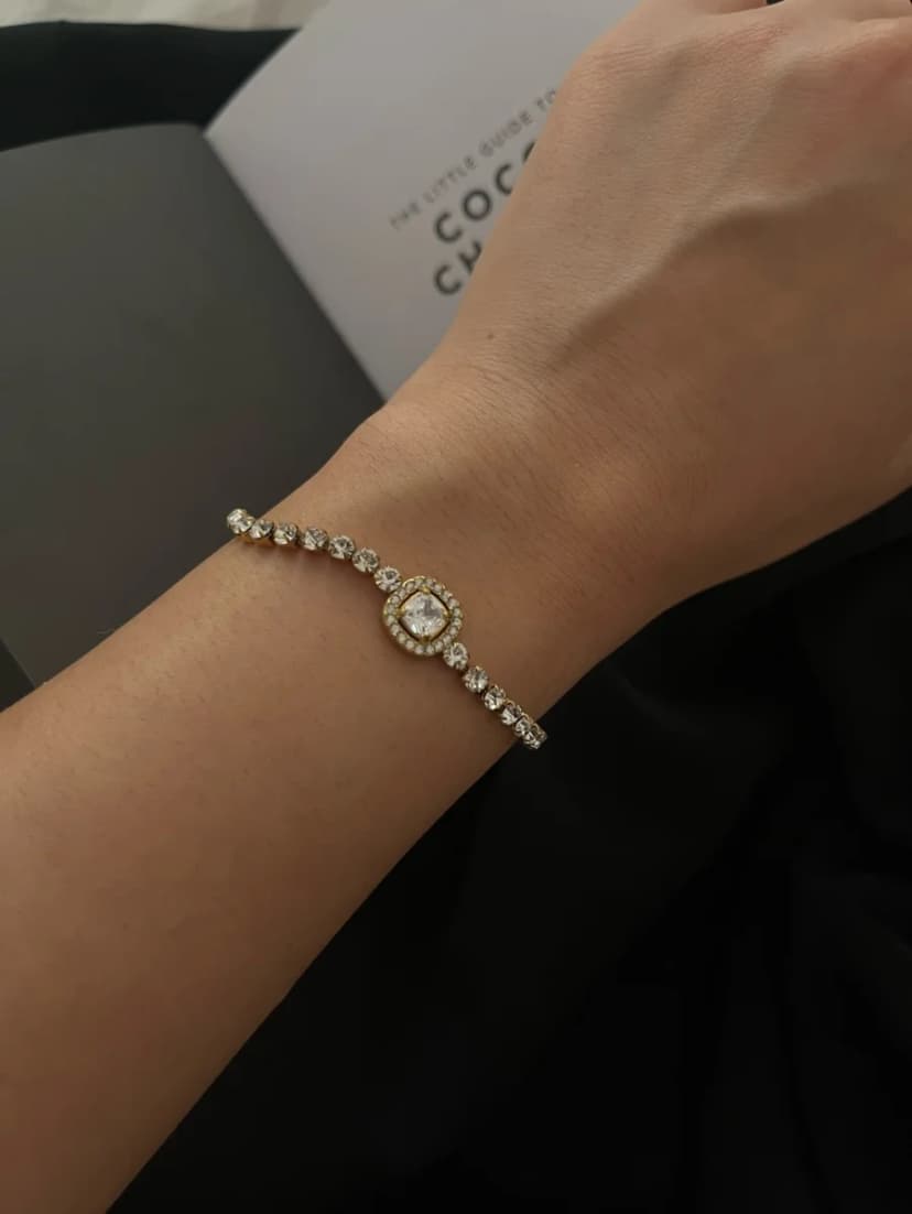 A person's wrist wearing a gold-tone bracelet with a central round, crystal-set halo charm and a crystal-studded chain. A dark background with a book