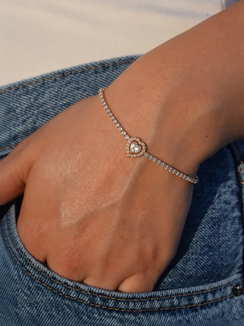 A person's wrist wearing a dainty gold-tone bracelet with a chain of small crystals and a sparkling heart charm.