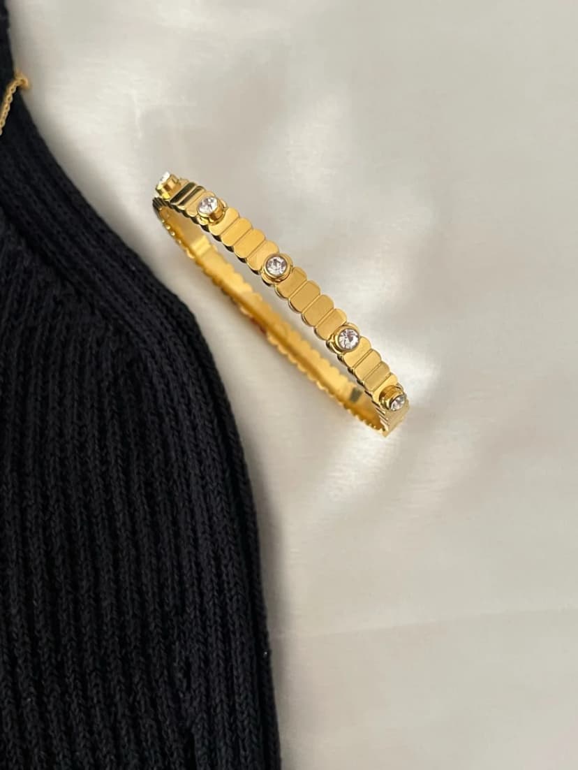A gold-tone bangle bracelet with embedded clear crystals rests on a light fabric next to a black knit top.