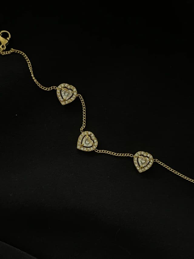 A gold-tone chain bracelet with three crystal-set heart charms is displayed on a black background.