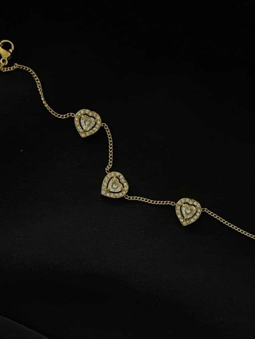 A gold-tone chain bracelet with three crystal-set heart charms is displayed on a black background.