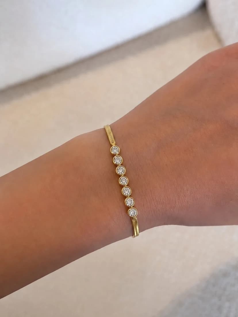 A person's light-skinned wrist wears a gold-tone flat snake chain bracelet with seven round, clear crystal-set stations.