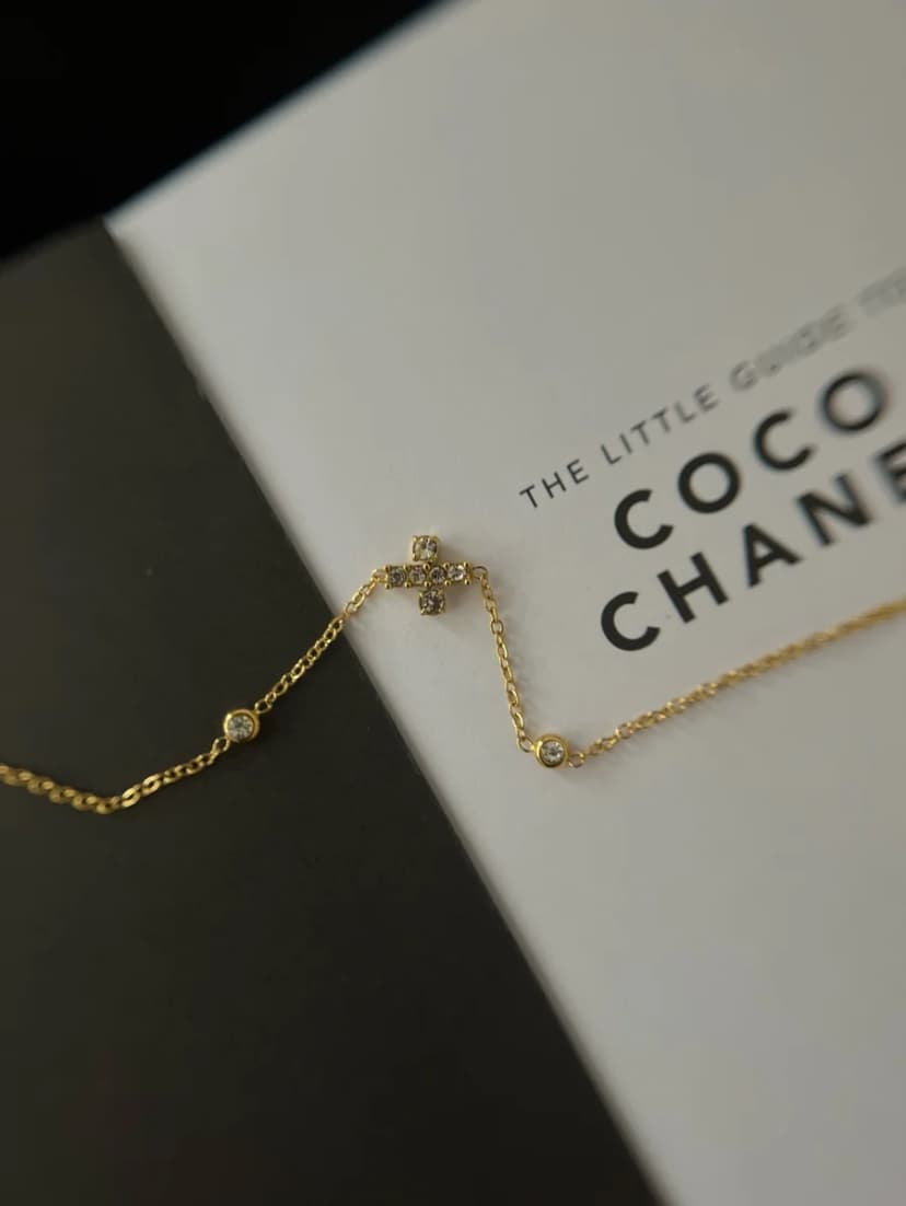 A fine gold-tone chain bracelet with a central crystal-set cross motif and two small crystal rounds, lying on an open book.