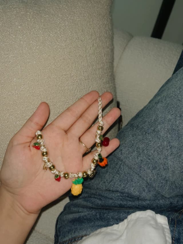 A person's hand holds a necklace with a beige braided cord, gold-tone beads, and colourful knitted fruit charms, resting on blue jeans.