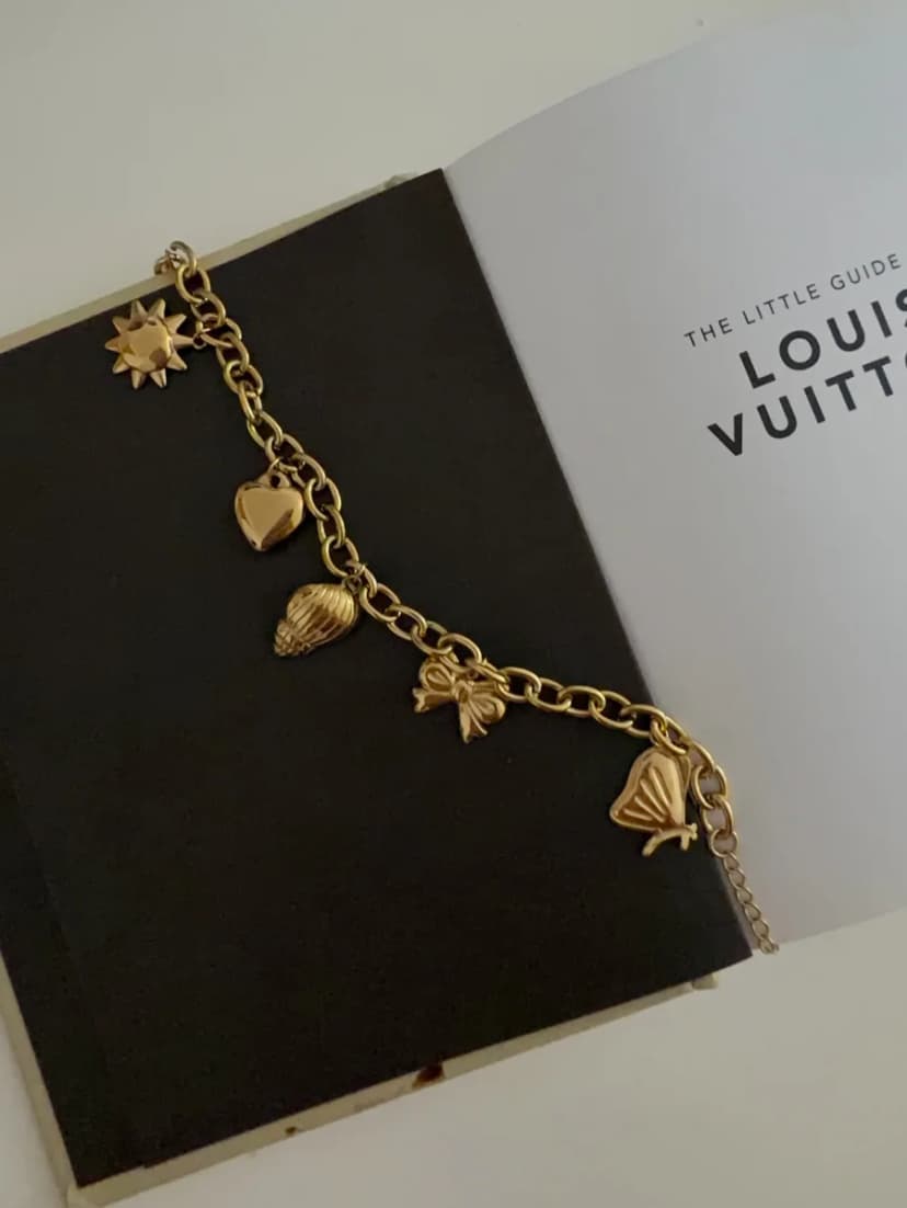 A gold-tone chain bracelet with sun, heart, shell, bow, and butterfly charms lies on a black book, next to a white page.