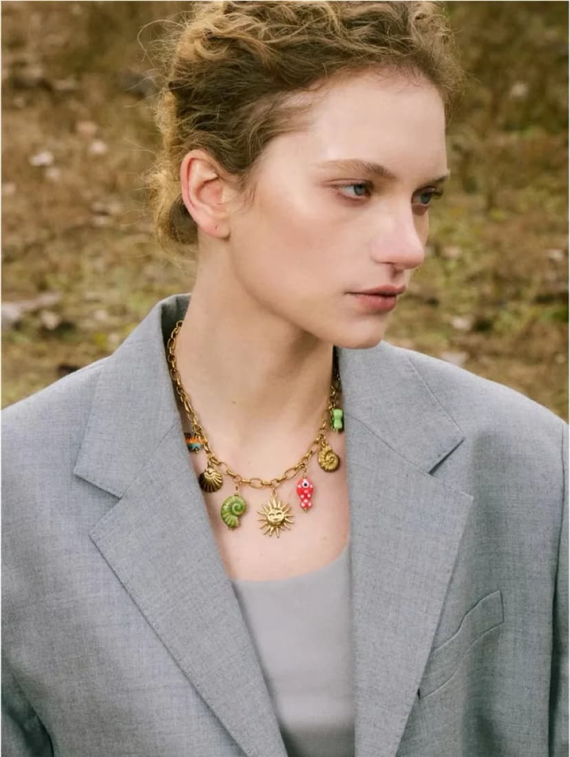 A woman with light brown hair wears a chunky gold-tone necklace with colourful sun, shell, and mushroom charms.