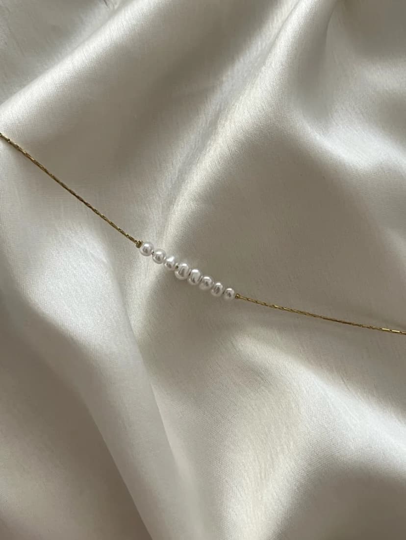 A dainty gold-tone chain bracelet with a row of seven small, round, white pearl-like beads, laid on a white satin fabric.
