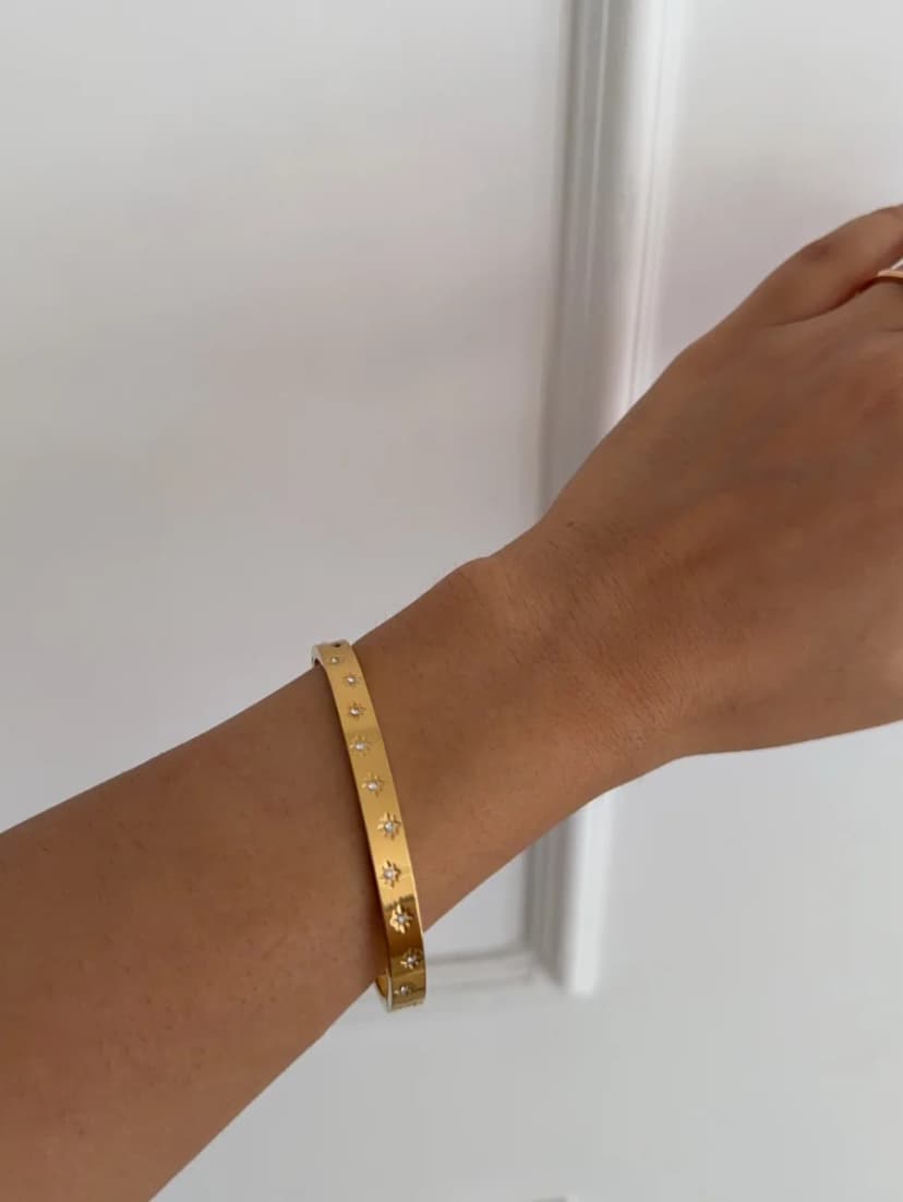 A medium-toned wrist wears a gold-tone cuff bracelet featuring small, clear, star-shaped crystal-like embellishments, against a white background.