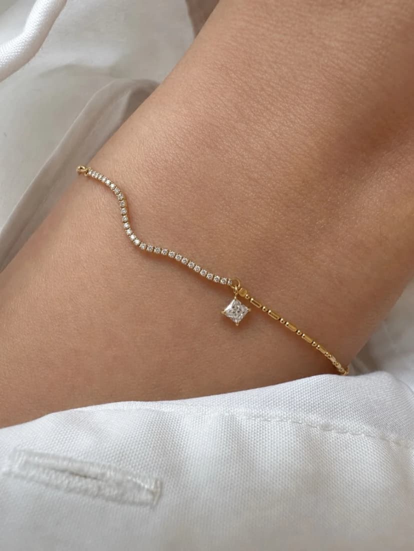A medium-toned arm wears a delicate gold-tone bracelet featuring a wavy line of tiny clear crystal-like stones and a single square crystal-like charm,