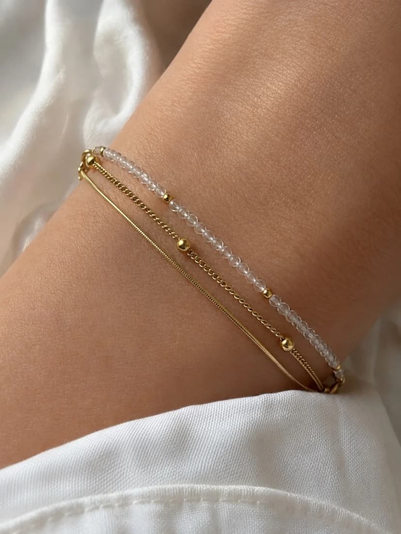 A medium-toned arm wears a layered gold-tone bracelet with strands of small clear beads, a fine chain, and tiny gold-tone beads, against white fabric.