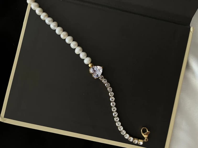 A bracelet with pearl-like beads, a crystal-set heart charm, and a gold-tone chain with clear crystal-like stones. It rests on a black book and white