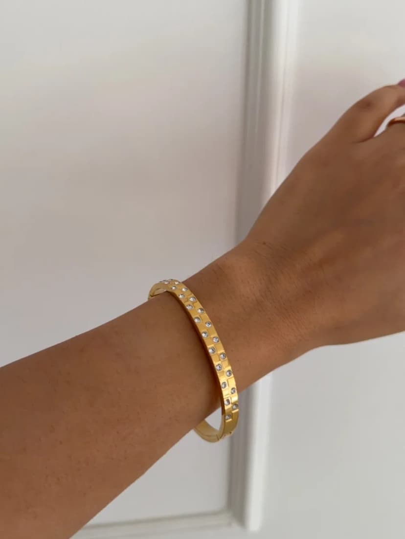 A medium-toned wrist wears a gold-tone cuff bracelet featuring small, clear, star-shaped crystal-like embellishments, against a white background.