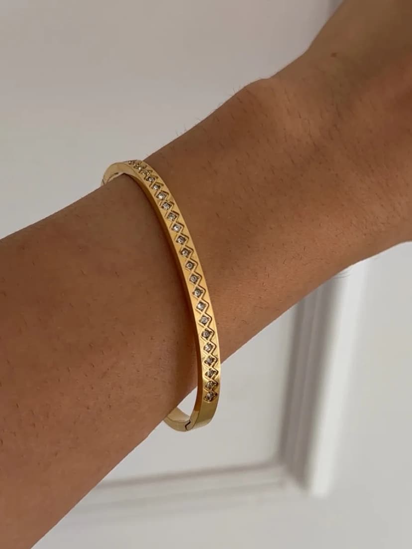 A gold-tone bangle bracelet with a pattern of clear crystal-like stones is worn on a person's wrist, against a white background.