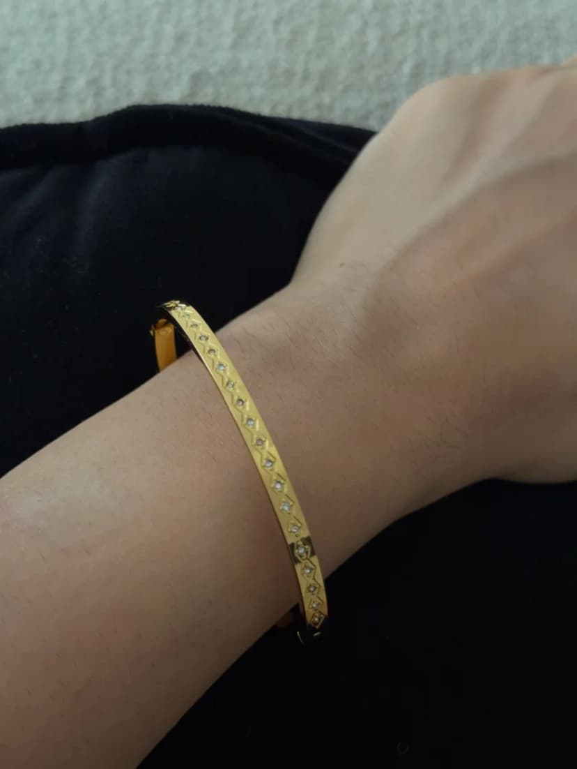A gold-tone bangle bracelet with a pattern of clear crystal-like stones is worn on a person's wrist, resting against dark, textured fabric.