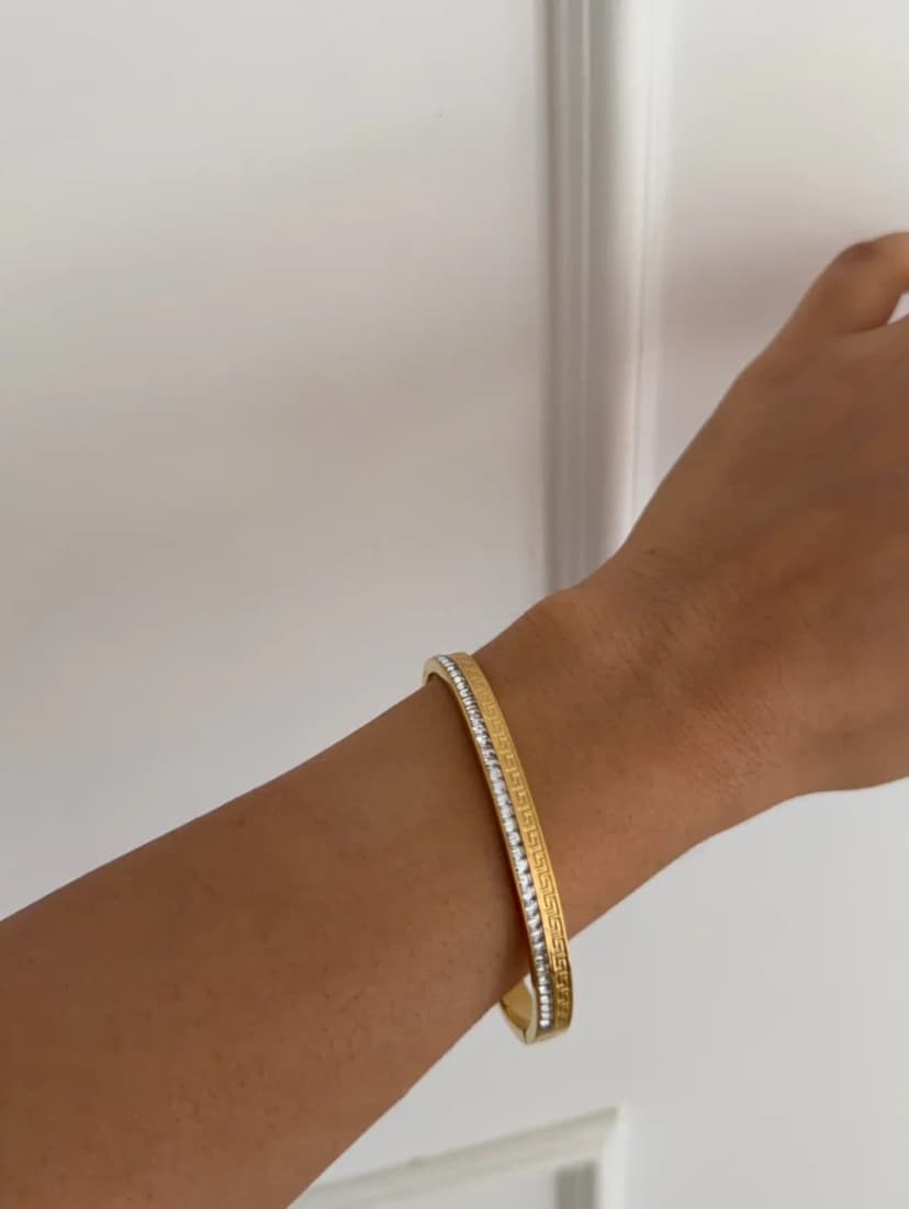 A gold-tone bangle bracelet with two bands, one textured and one set with clear crystal-like stones, worn on a person's wrist against a white door.