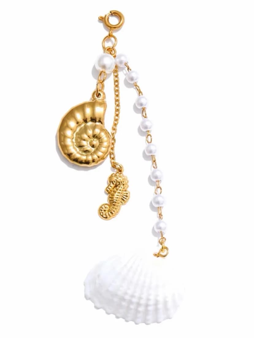 A gold-tone charm with a spiral shell, a seahorse, and a strand of pearl-like beads, displayed next to a white seashell on a white background.