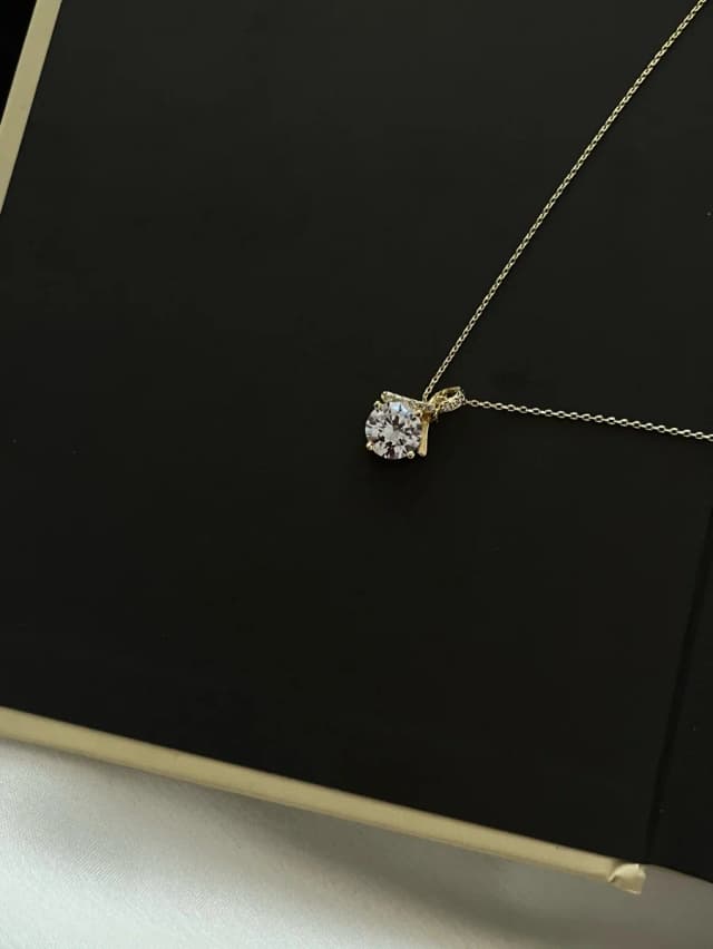 A delicate gold-tone necklace with a single large, round, clear crystal pendant in a four-prong setting, displayed on a black surface.