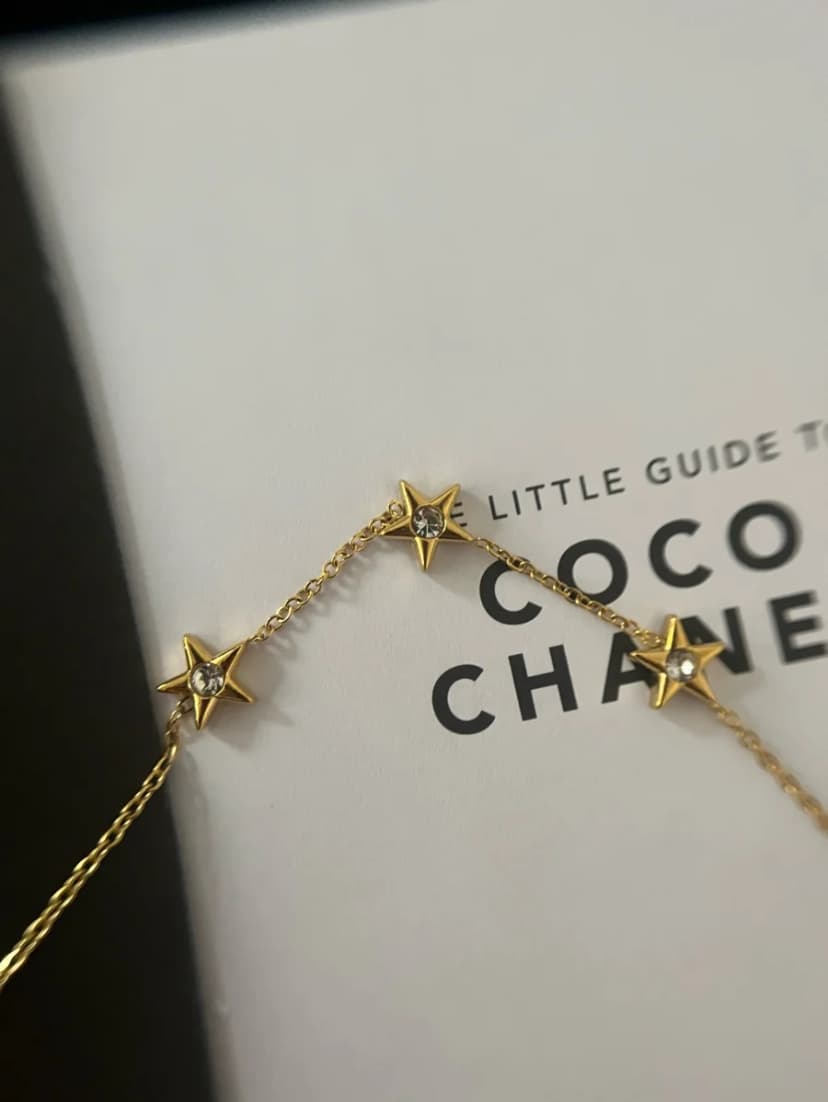 A delicate gold-tone chain bracelet with three crystal-set star charms, draped over a white book titled "THE LITTLE GUIDE TO COCO CHANEL".