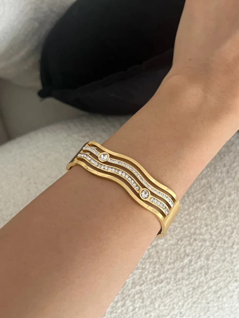 A wide gold-tone cuff bracelet with three wavy bands, featuring small and larger clear crystals, worn on a person's tanned wrist.