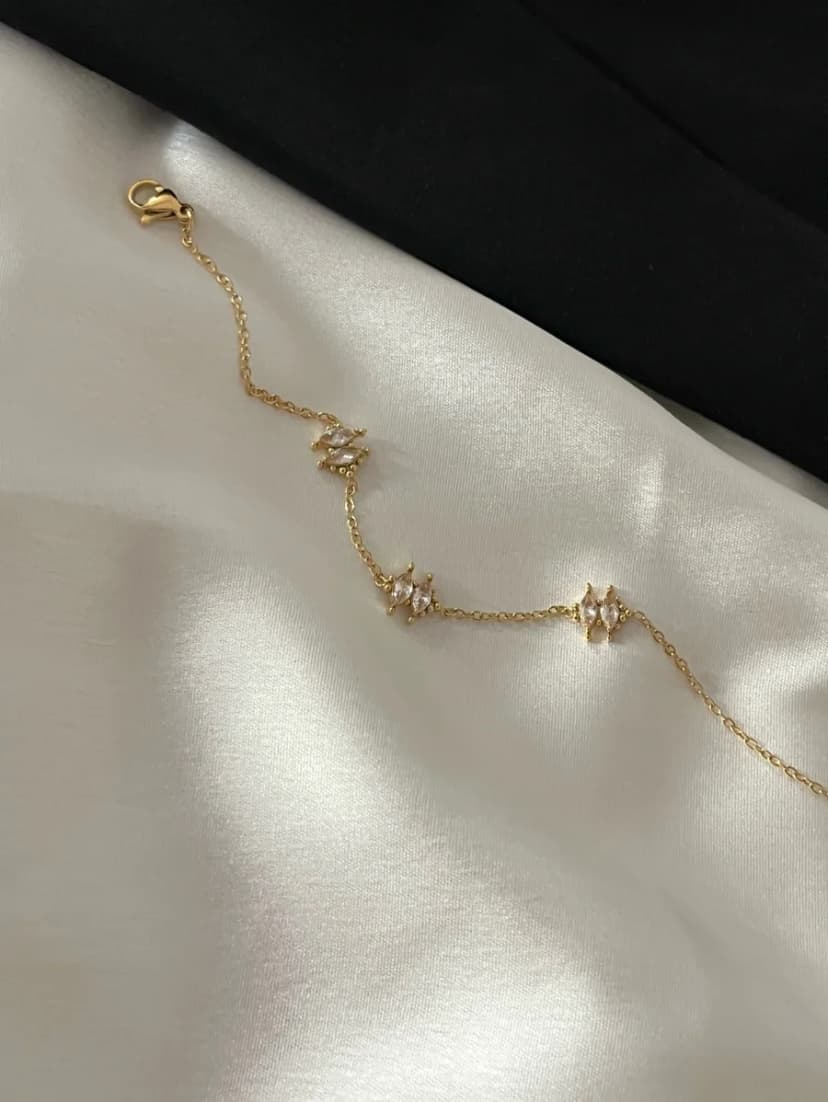 A delicate gold-tone chain bracelet with three evenly spaced, pale pink crystal-set charms, displayed on white satin fabric.