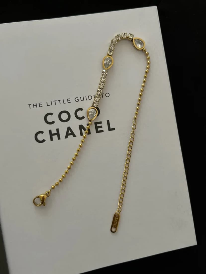 A gold-tone bracelet with a line of clear crystals, three teardrop-shaped crystal-set motifs, and a fine bead chain, on a white book.
