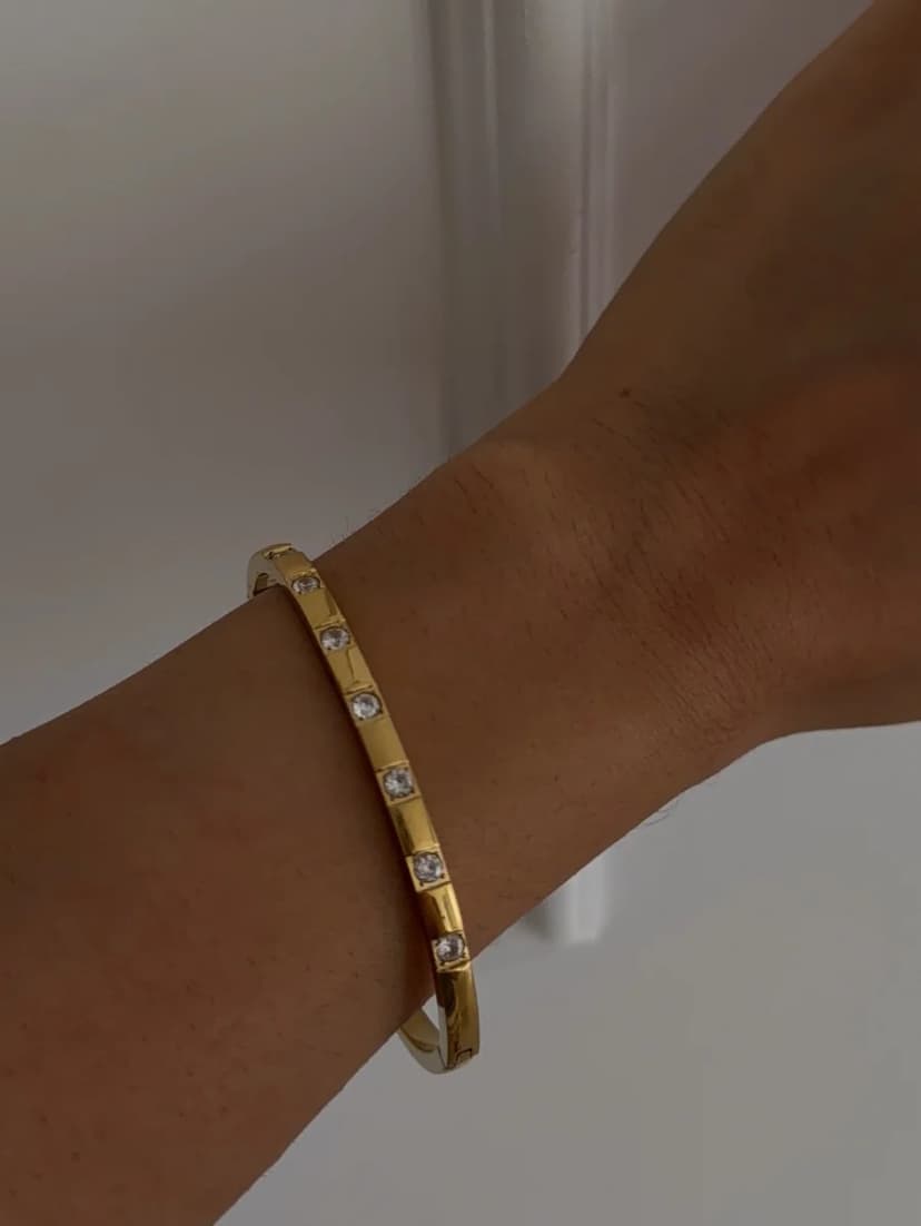A gold-tone bangle bracelet with clear square-cut stones is worn on a person's wrist against a light background.