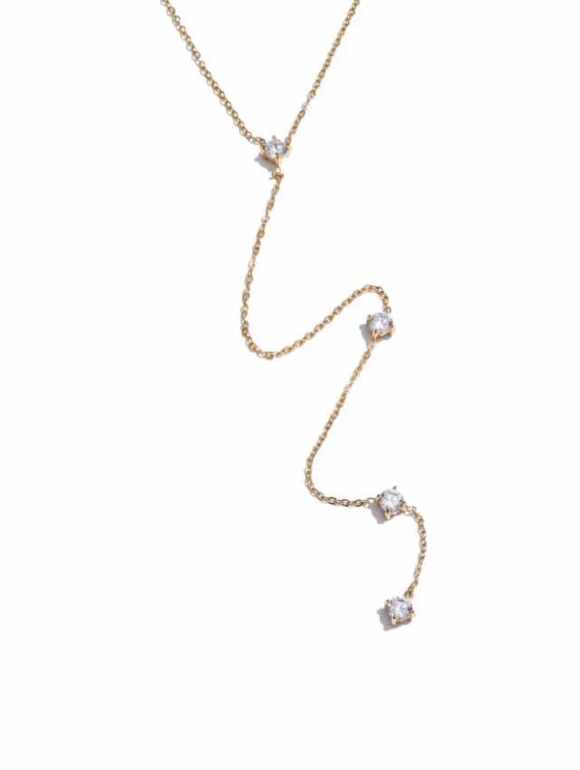A delicate gold-tone lariat necklace with four individual round clear crystal-like stones spaced along its drop on a white background.